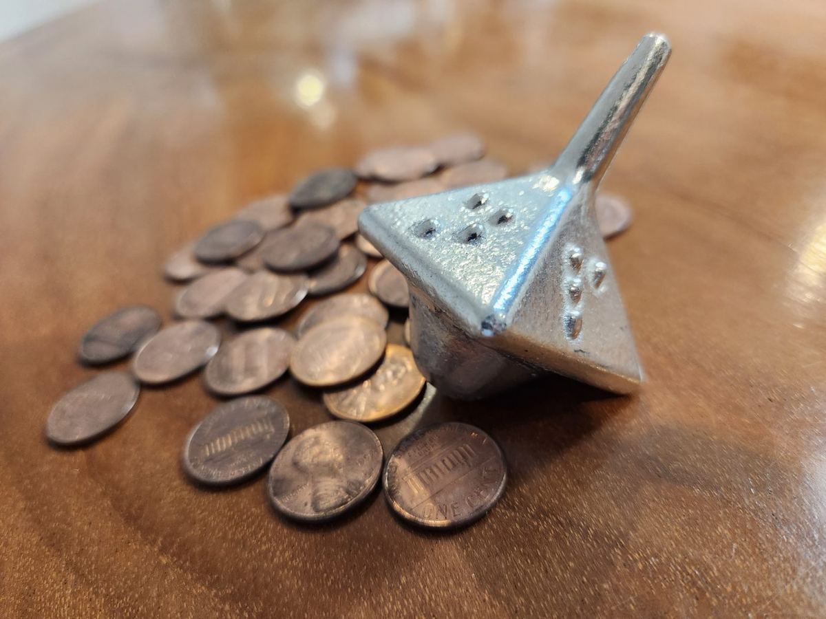 A silver dreidel with raised Braille dots on its side, resting beside a scattered pile of pennies.
