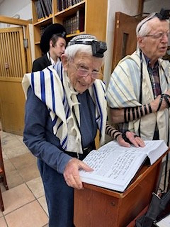 An older man smiling as he prays, wrapped in a tallis and wearing tefillin, one hand resting on one of our open large print books, with other men praying beside him.