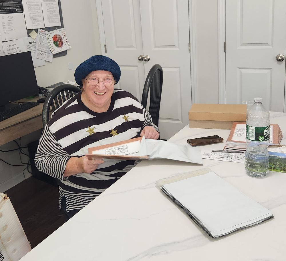 Mrs. Lena Cohen, smiling, sits at a table holding a large calendar and a white mailing envelope, with more envelopes and calendars spread out in front of her.