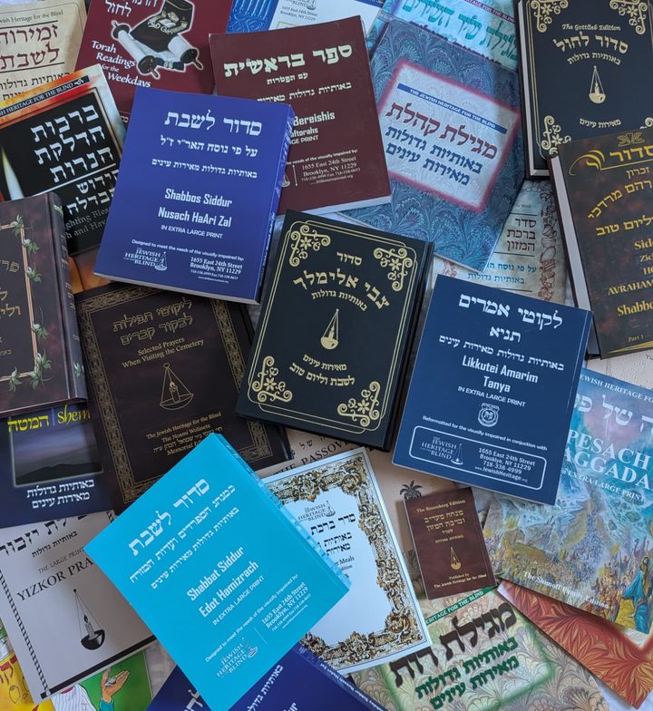 A table covered with dozens of our large print books: Siddurim for Shabbos and weekdays in Nusach HaAri and Edot Hamizrach, Tanya, Sefer Bereishis, Megillas Koheles and Megillas Rus, a Pesach Haggadah, Zemiros, Birchas Hamazon, Yizkor prayers, Selected Prayers When Visiting the Cemetery, and a children’s storybook.