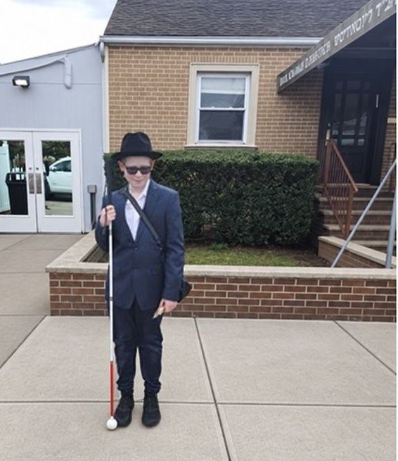 A boy in a dark suit, black hat and sunglasses stands on a sidewalk holding a white cane, in front of a brick building with a hedge.
